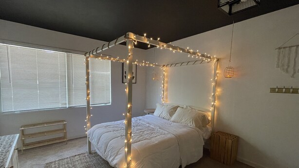 Un dormitorio con cama con dosel, una mesita de noche, una alfombra pequeña y una ventana con persianas.