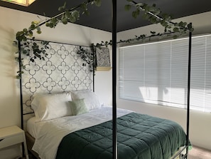 Cama com colcha verde, travesseiros brancos e cabeceira de metal preta com padrão geométrico.