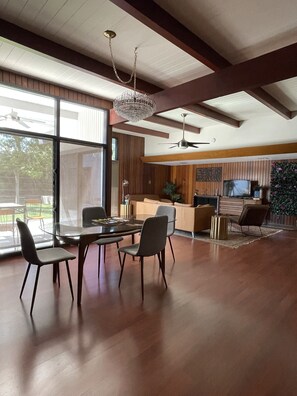Uma sala de estar ampla com mesa de jantar de vidro, cadeiras modernas, ventilador de teto e uma janela grande.
