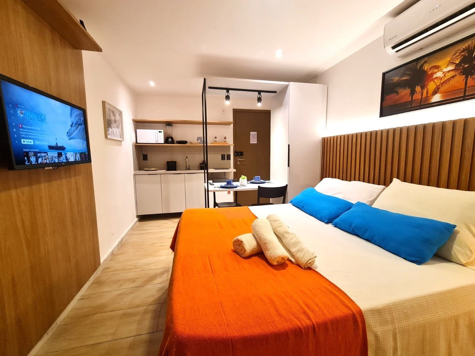 A hotel room with a bed, a television, a kitchenette, and a painting on the wall.