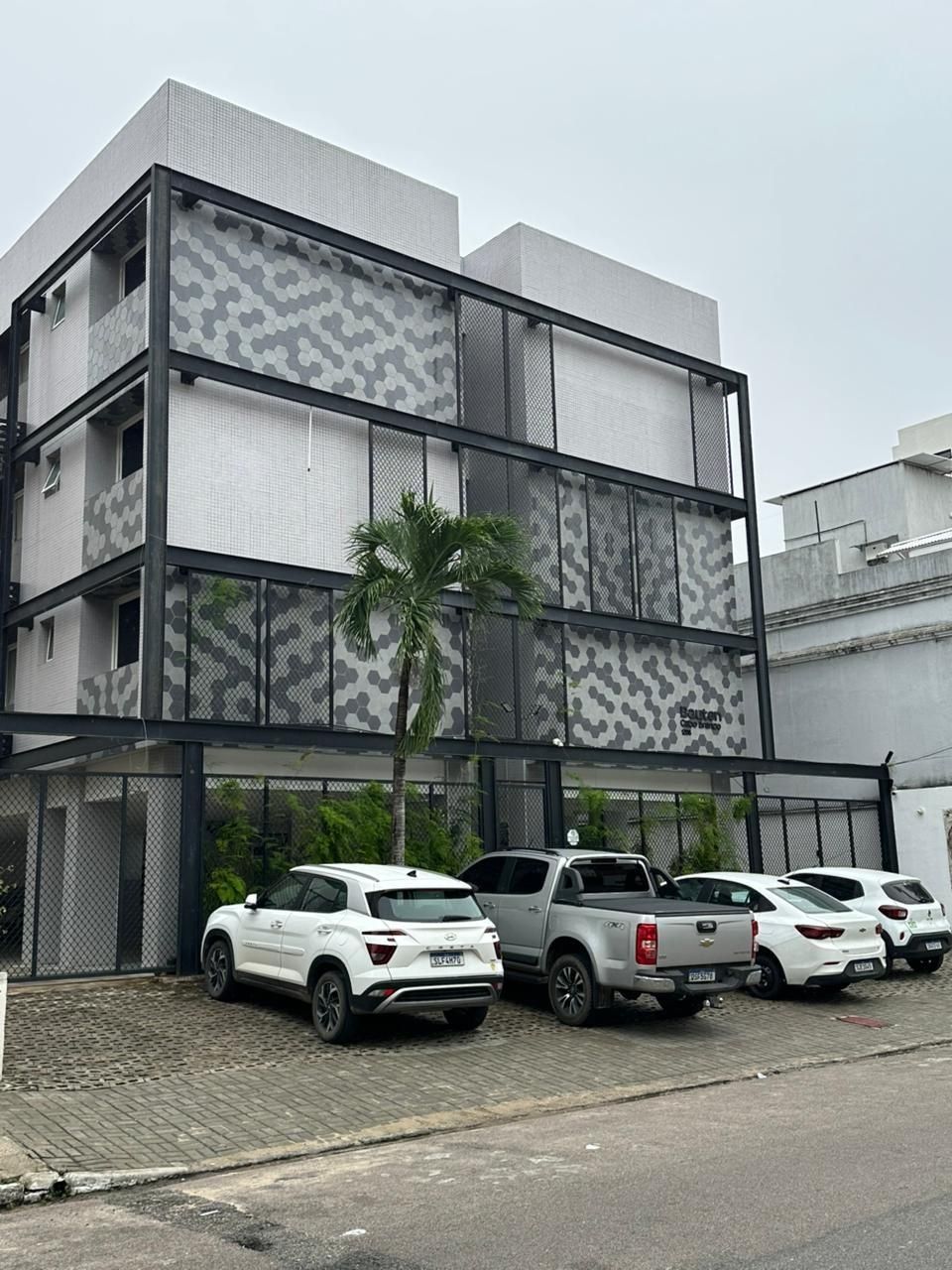 Un edificio de varios pisos con fachada ornamentada, una zona de estacionamiento con varios vehículos y una palmera frente a él.