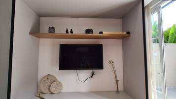 Una habitación con televisor de pantalla plana montado, un estante de madera con adornos, un enchufe en la pared y una ventana con vistas a zonas verdes.