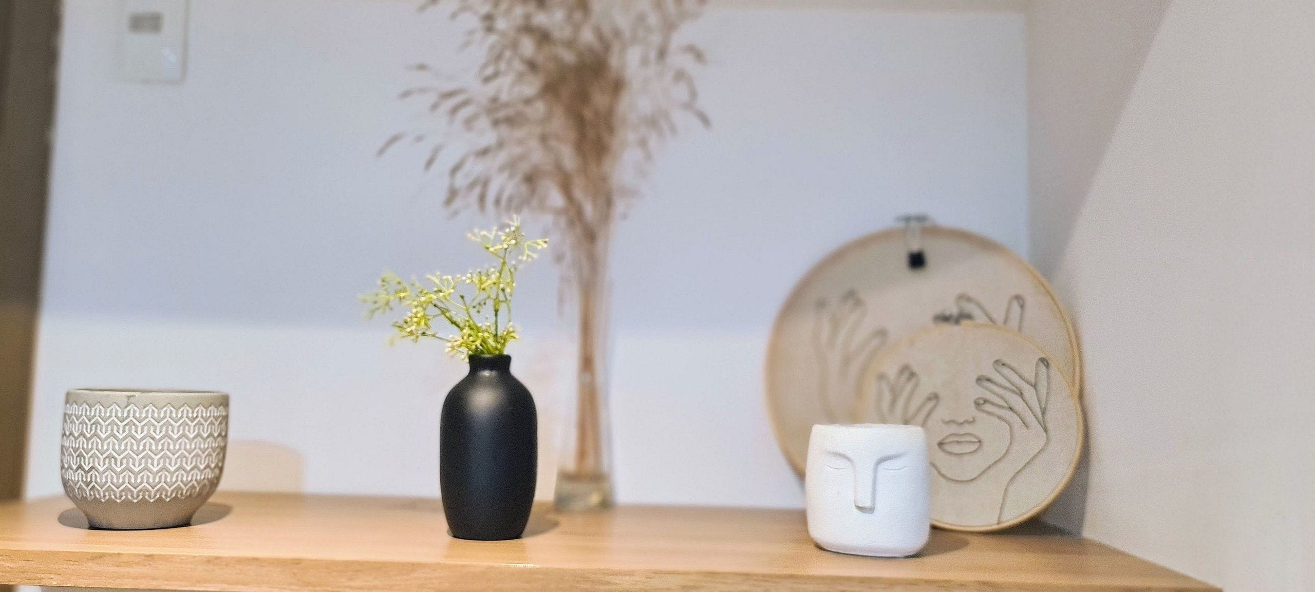 A shelf with three decorative items: a patterned ceramic cup, a black vase with flowers, and a white ceramic mug.