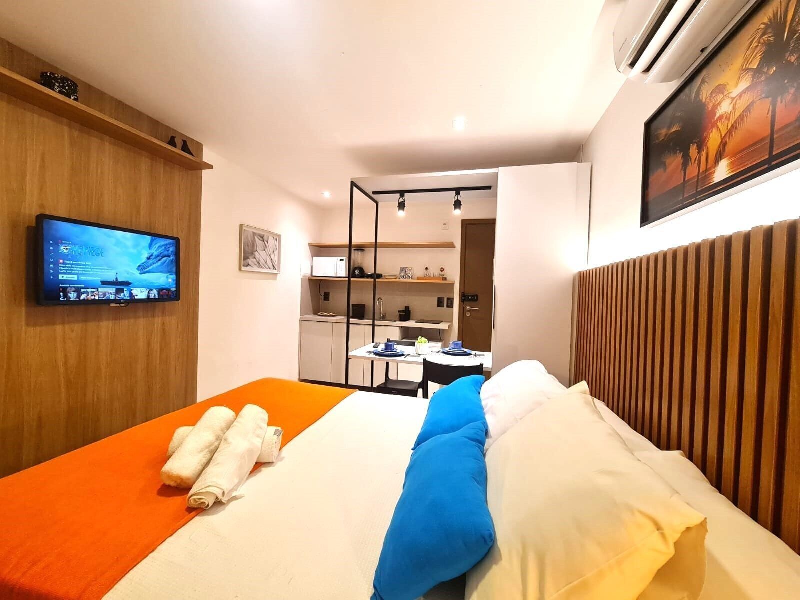 A hotel room with a bed, a television, a kitchenette, and a painting on the wall.