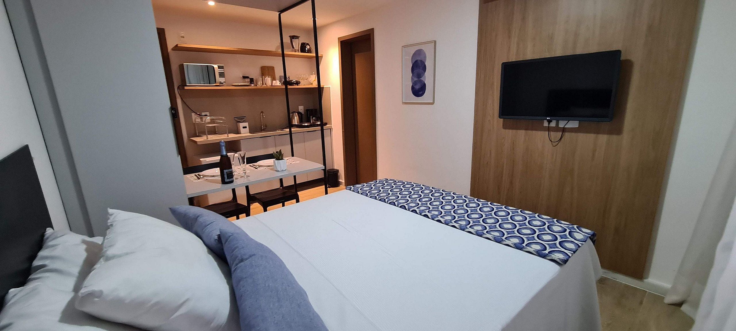 Un dormitorio con cama, televisor montado en la pared y una pequeña cocina con estantes.