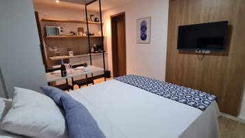 Un dormitorio con cama, televisor montado en la pared y una pequeña cocina con estantes.