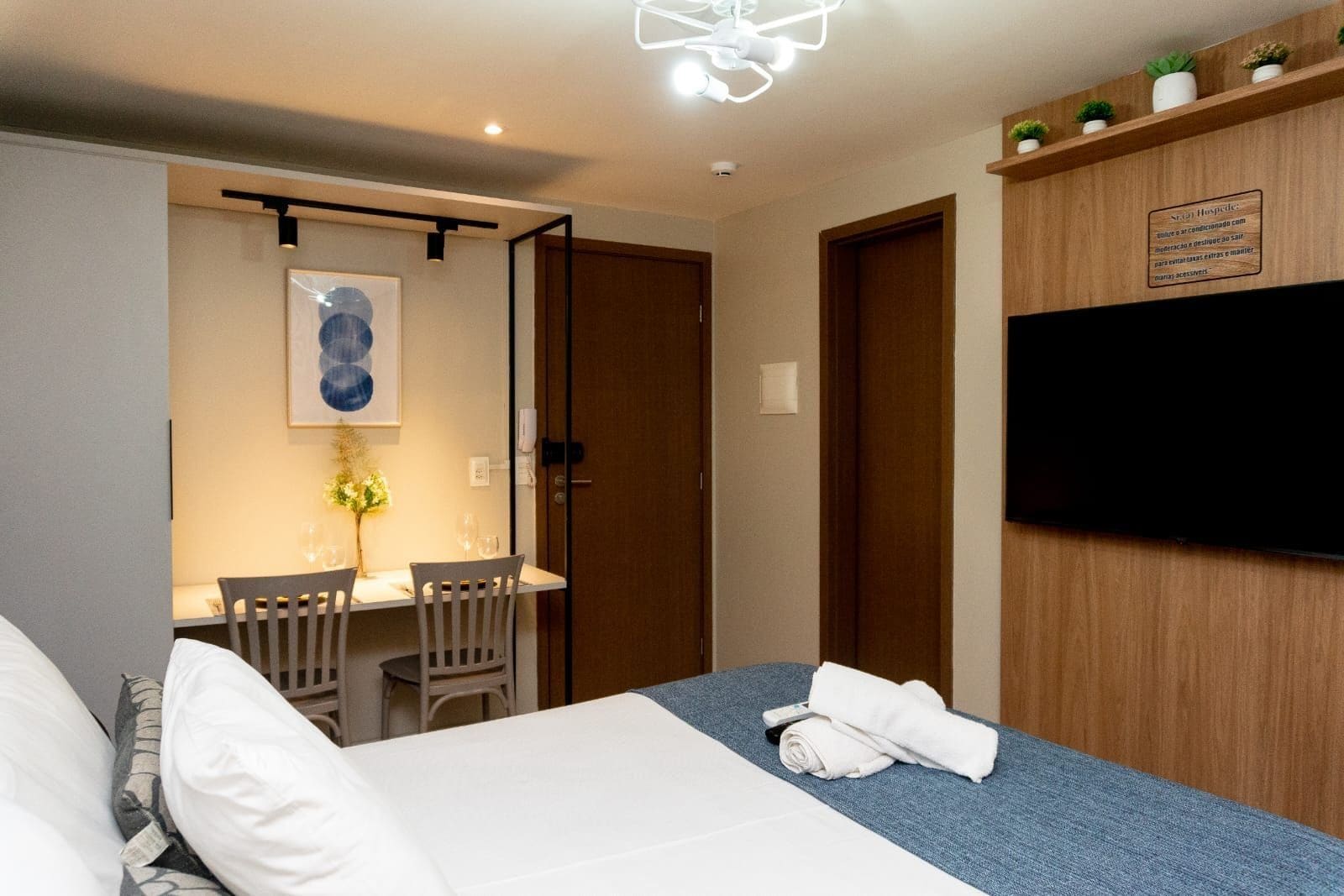 A hotel room with a bed, a desk with a chair, a television, and a small shelf with plants.