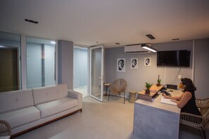 Una sala moderna con un sofá, un comedor con mesa y sillas, un televisor montado en la pared y un puerta de vidrio que da a otra habitación.