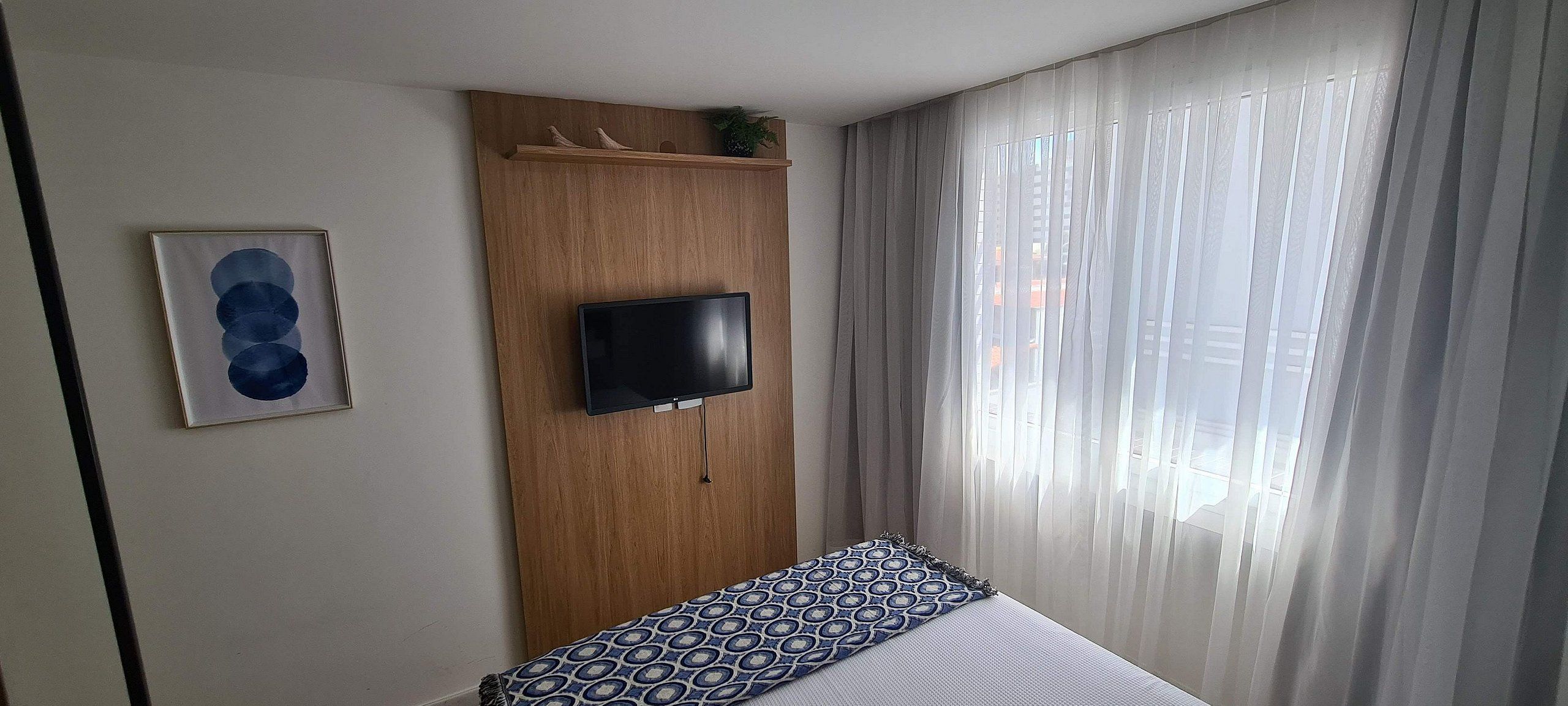 Ein Hotelzimmer mit einem Bett, einem an der Wand befestigten Fernseher, einem gerahmten Kunstwerk und einem Fenster mit durchsichtigen Vorhängen.