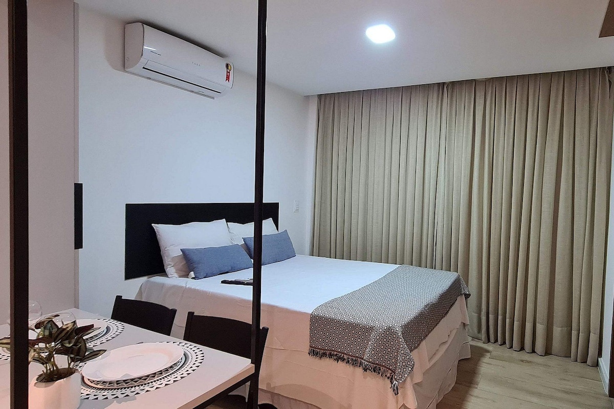Habitación de hotel con una cama, televisor montado en la pared, una mesa de comedor con una planta y una ventana con cortinas.