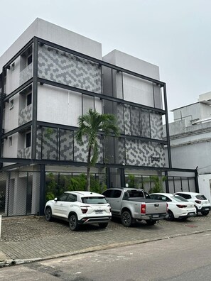 Un edificio de varios pisos con fachada ornamentada, una zona de estacionamiento con varios vehículos y una palmera frente a él.