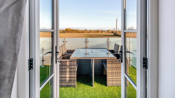Une véranda fermée par des baies vitrées, avec une table et des chaises, offrant une vue sur un champ verdoyant et un paysage lointain.