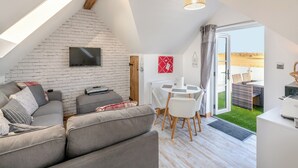 Un espace de vie moderne de style loft, avec un plafond en pente, un téléviseur à écran plat fixé sur un mur en briques blanches, un canapé gris agrémenté de coussins à motifs, un coin repas avec une table et des chaises blanches, et une vue sur un champ verdoyant par une porte vitrée.