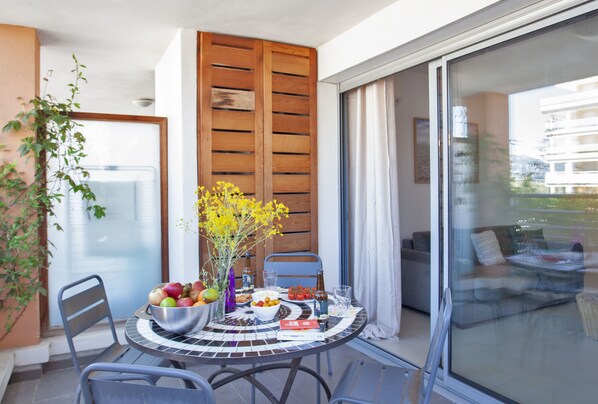 A dining area with a round table, chairs, a bowl of fruit, a vase with flowers, and a view of the outside through a sliding glass door. - Appartement "Fiora" en centre-ville de Calvi - 2 4 personnes (Calvi)