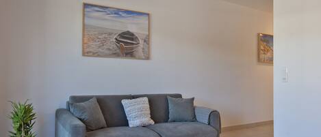 Una sala moderna con un sofá gris, un cuadro de un barco sobre la arena y una planta pequeña.