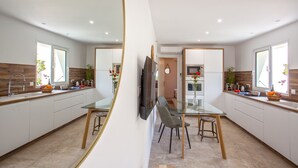 A modern kitchen with white cabinets, a glass dining table, and a TV mounted on the wall. - Appartement neuf "Magnolia", terrasse pergola, jardin privatif - 2 4 pers (Calvi)