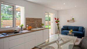 A modern kitchen with a glass dining table, white cabinets, and a blue sofa in the living area. - Appartement neuf "Magnolia", terrasse pergola, jardin privatif - 2 4 pers (Calvi)