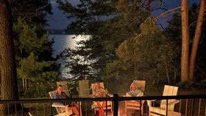 Eine Terrasse mit Adirondack-Stühlen mit Blick auf einen See bei Nacht, in der Mitte brennt ein Feuer.