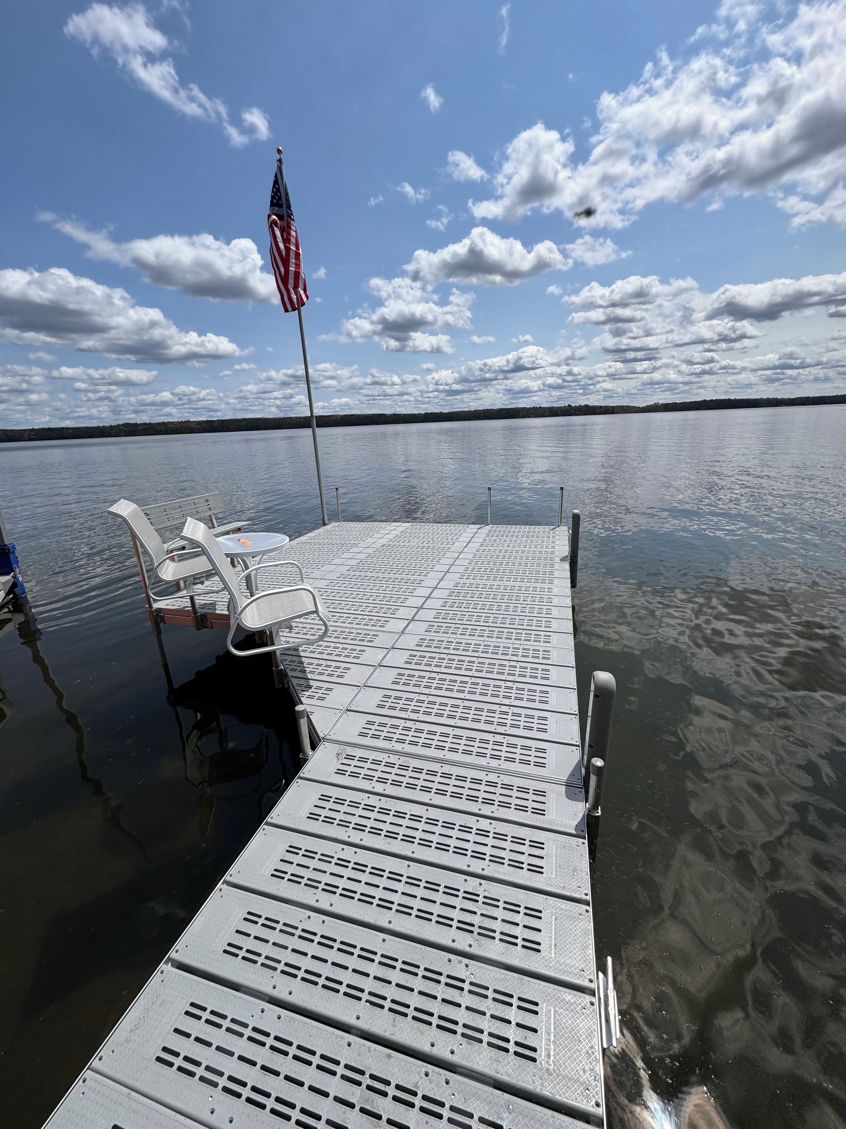 A dock with metal grates and white plastic chairs, an American flag, and calm water under a partly cloudy sky.