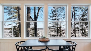Ein Essbereich mit Tisch und Stühlen, große Fenster mit Blick auf Bäume und ein Gewässer.