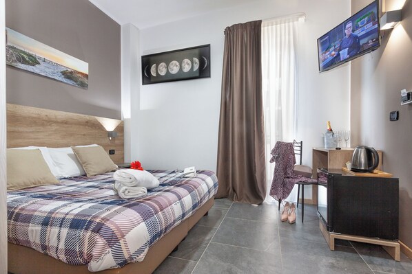 Quarto de hotel com cama, escrivaninha, cadeira, televisão e janela com cortinas.