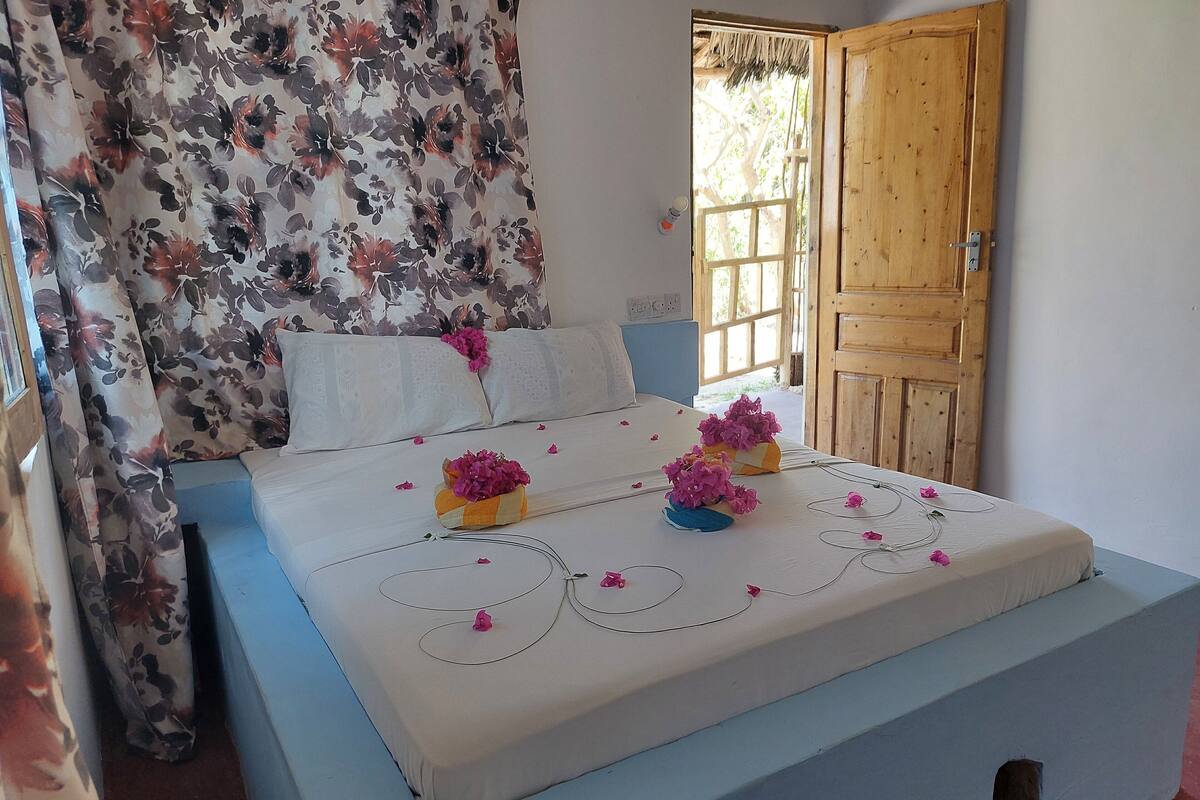 Un llit ben fet amb cortines de motius florals, una porta de fusta i vista a l'exterior.
