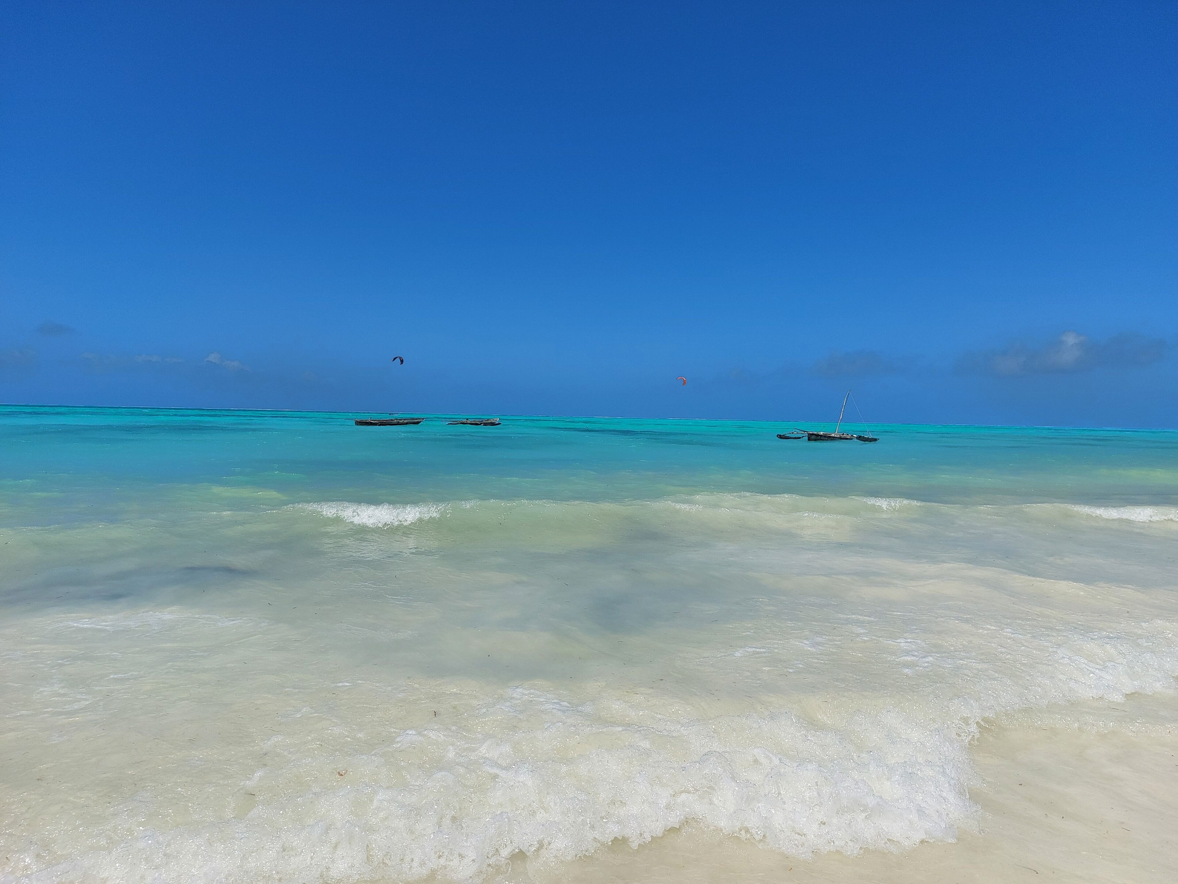 A beach with clear turquoise water, calm waves, and a few boats anchored in the distance.