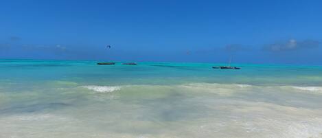 A beach with clear turquoise water, calm waves, and a few boats anchored in the distance.