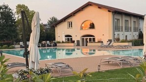 Un área de piscina con sillones de descanso y sombrillas, un edificio grande con ventanales arqueados y un césped bien cuidado.