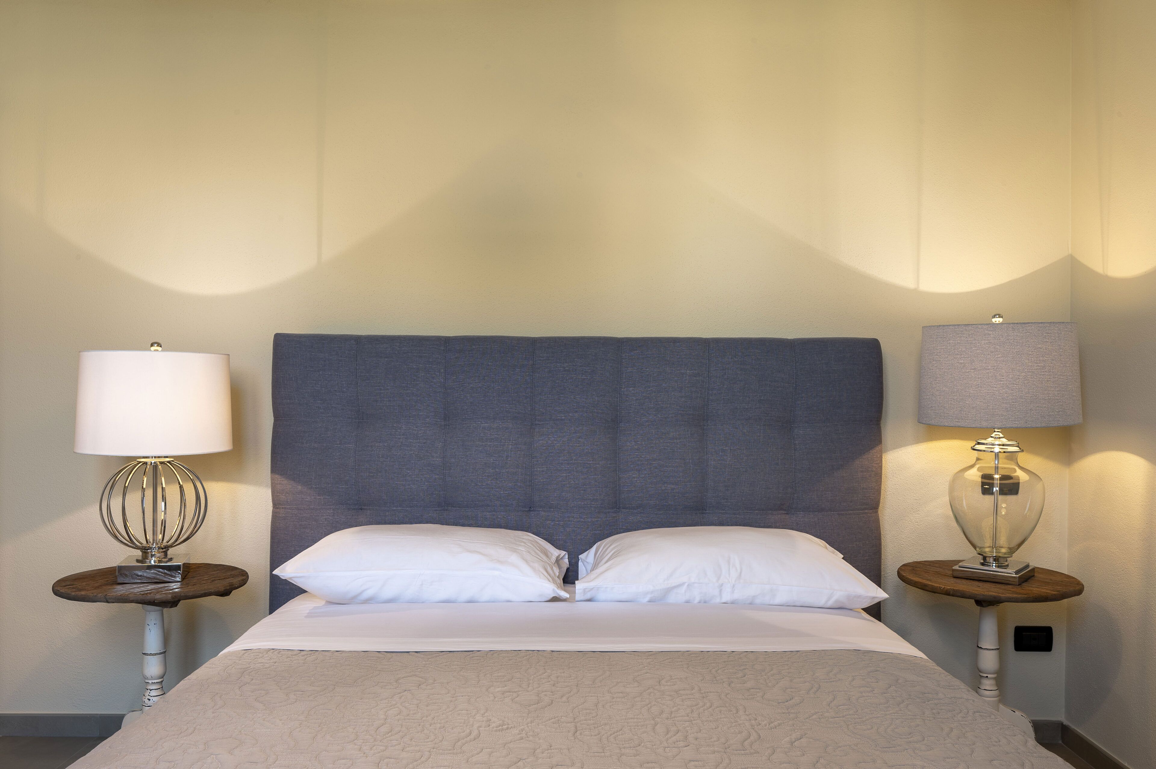 A neatly made bed with two pillows, flanked by two bedside tables with lamps, in a room with a plain wall and a large, padded headboard.