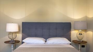 A neatly made bed with two pillows, flanked by two bedside tables with lamps, in a room with a plain wall and a large, padded headboard.