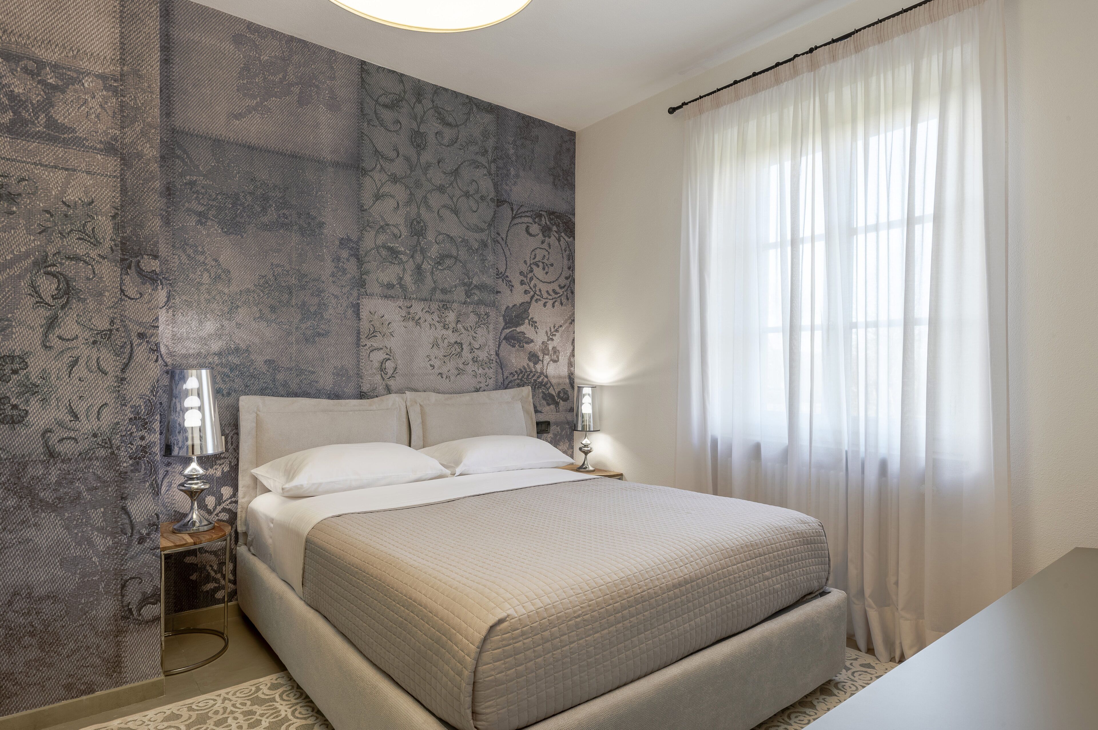Una cama bien hecha en una habitación con papel tapiz estampado, una lámpara de noche y una ventana con cortinas traslúcidas.