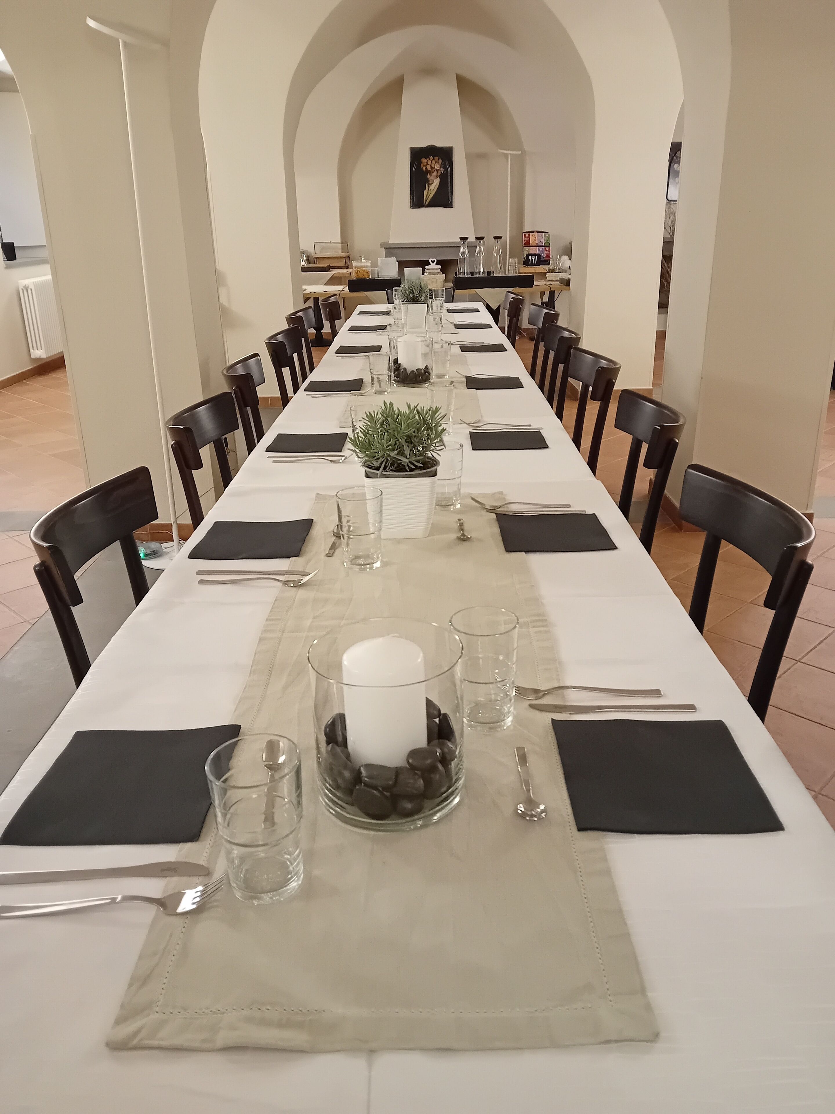 Una larga mesa de comedor con sillas negras, mantel blanco y centros de mesa que incluyen una vela y una planta en maceta.