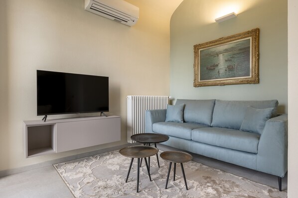 A modern living room with a flat-screen TV, a grey sofa, two round side tables, and a patterned rug. - Villa Raggi Relais (Castelletto d'Orba)