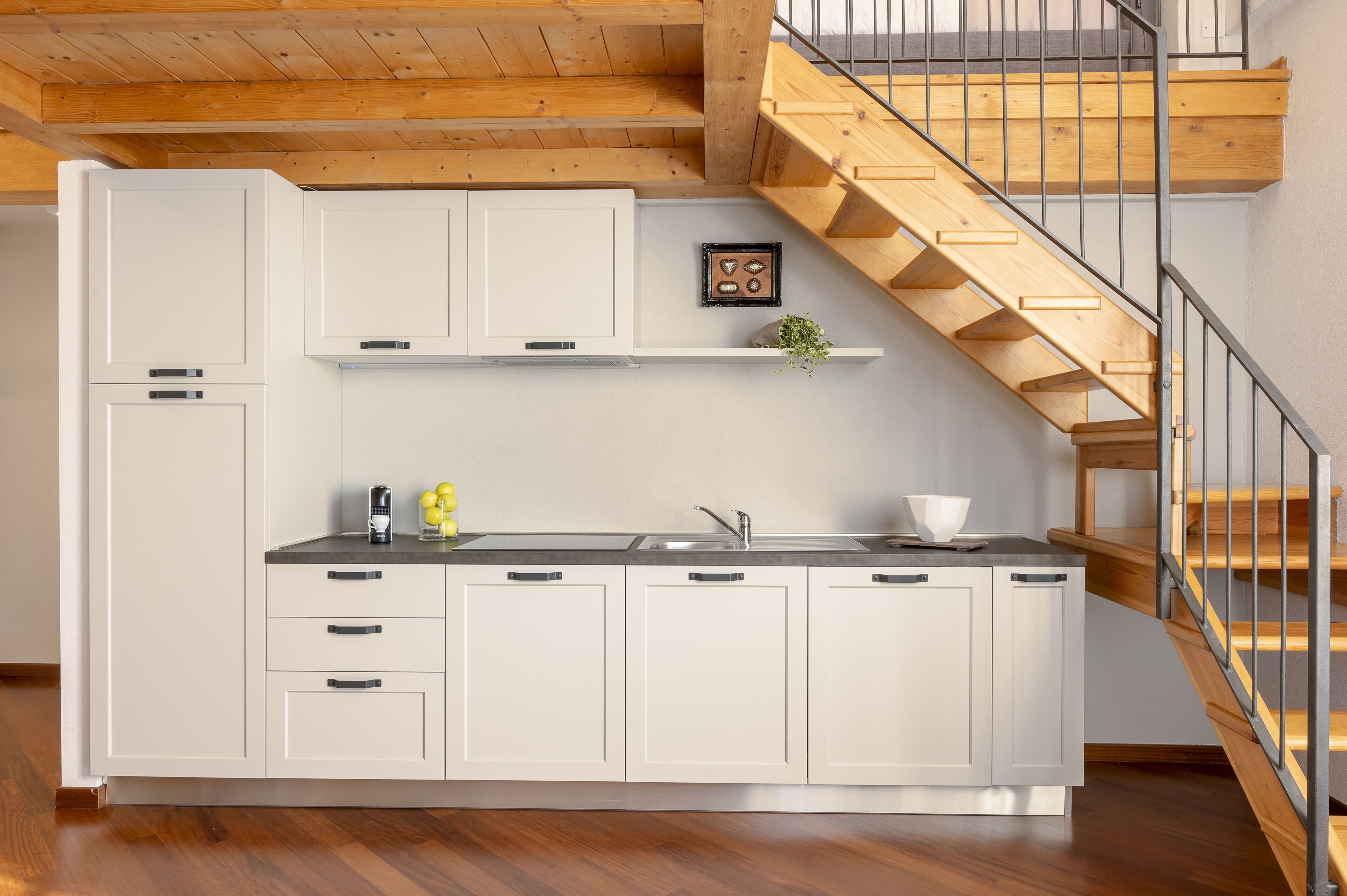 A modern kitchen with white cabinets, a wooden staircase, and a small plant on the shelf.
