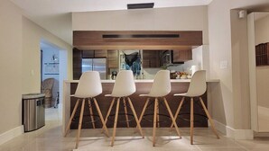 A modern kitchen with a central island, four white bar stools, and a trash bin.