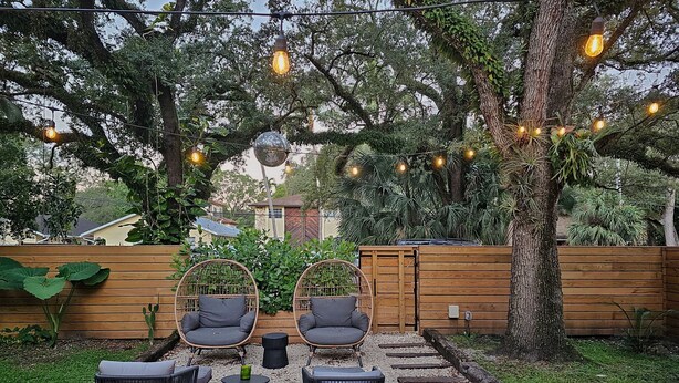 An outdoor seating area with wicker chairs and a small table, surrounded by trees and a wooden fence.