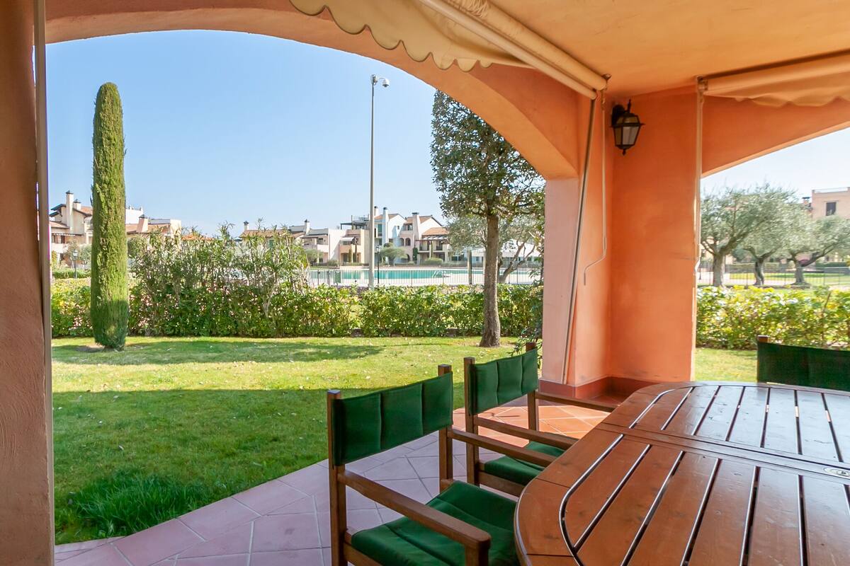 Un pati exterior amb taula de fusta i cadires amb coixins verds, amb vista a un jardí ben cuidat i edificis allunyats.
