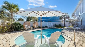 Una piscina con dos sillas Adirondack bajo un paraguas grande, rodeada de un patio de ladrillo y una valla blanca.