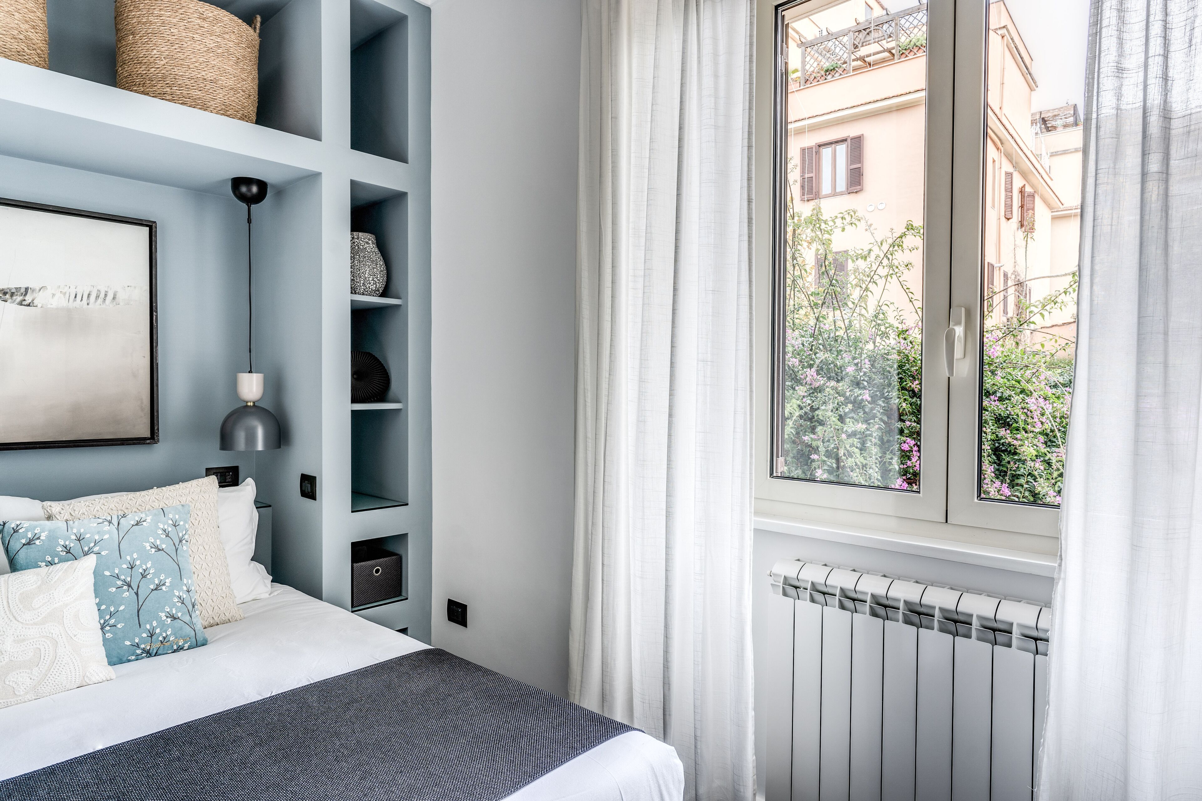 Une chambre à coucher avec un lit, une fenêtre avec des rideaux, un radiateur et une vue sur les bâtiments à l’extérieur.