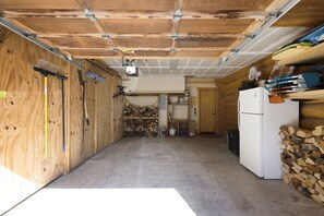 Un garage avec un sol en béton, des murs en bois, un réfrigérateur blanc et des étagères remplies d’outils et de bois.
