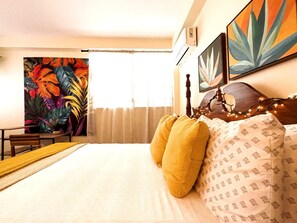Una habitación de hotel con una cama, cabecera de madera, un cuadro de hojas tropicales y una ventana con cortinas.