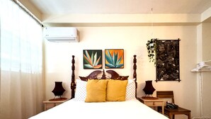 A neatly made bed with a wooden headboard, two wall-mounted paintings, a small table with a lamp, and a hanging plant. - Charming Boho 2 bedroom in Miramar - Back 2B (San Juan)