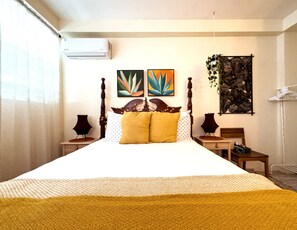 Una cama bien hecha con cabecera de madera, dos cuadros colgados en la pared, una mesita con una lámpara y una planta colgante.