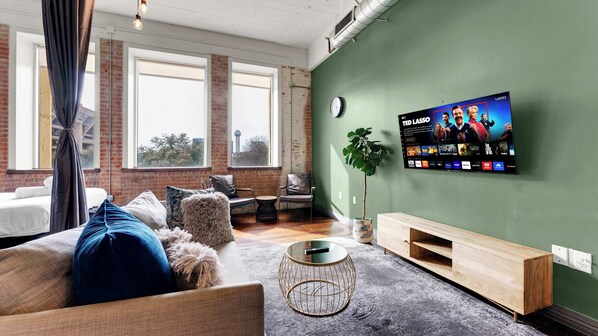 Un salon moderne doté d’un grand téléviseur à écran plat fixé au mur, d’un coin salon confortable avec un canapé et des fauteuils, d’une petite table basse ronde et d’une grande fenêtre laissant entrer la lumière naturelle.