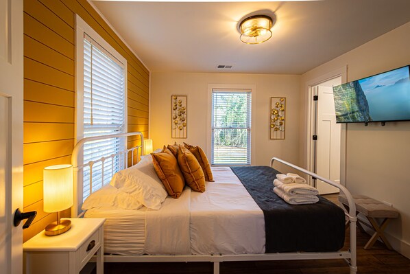 A bedroom with a bed, bedside lamp, window blinds, a television, and a small bench.