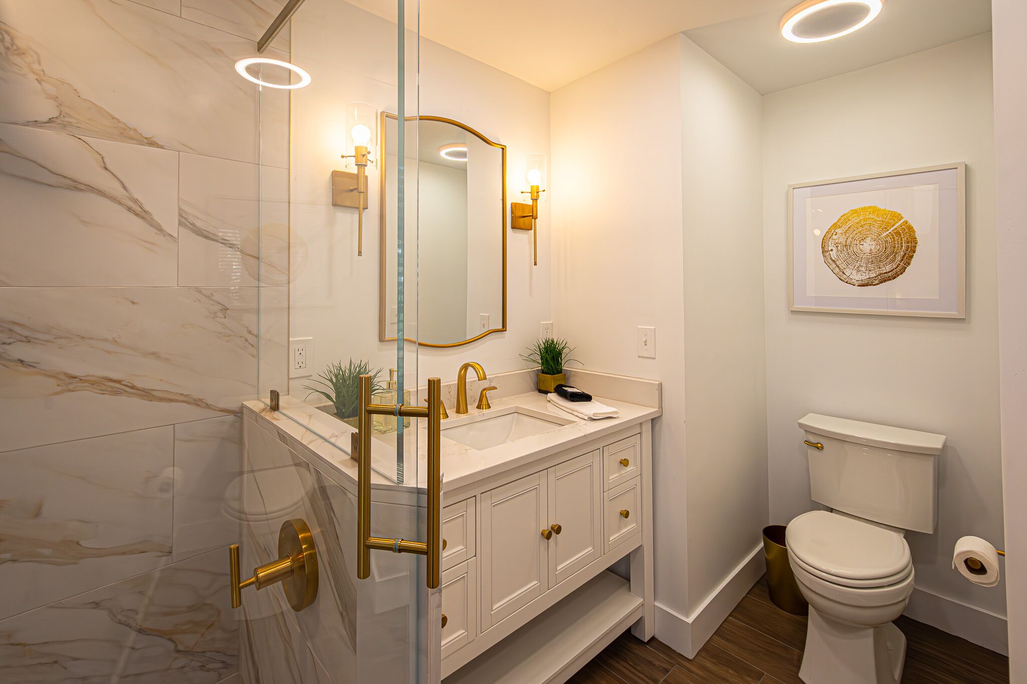 Une salle de bain moderne avec une douche à l’italienne en verre, un meuble-lavabo blanc agrémenté de robinetterie dorée et une œuvre d’art encadrée au mur.