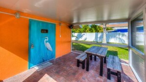 Un porche cubierto con una puerta azul decorada con un mural de pájaros, una mesa de madera y un banco.