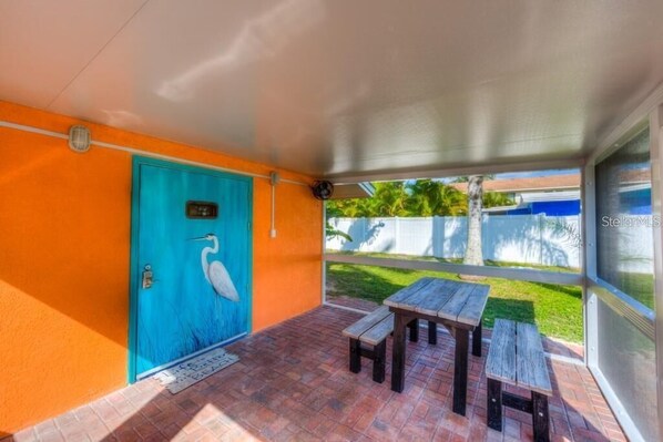 Un porche cubierto con una puerta azul decorada con un mural de pájaros, una mesa de madera y un banco.