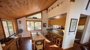 A cozy cabin interior with a wooden dining table, bar stools, a kitchen area, and a large window providing natural light. - Stunning 3-Bed 2 Bath Cabin with Loft @ Northstar (Truckee)